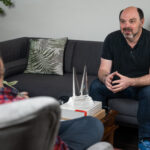 Morris Misel seated at a desk, holding a smartphone displaying the word “AI,” with a laptop beside him showing the word “think,” symbolising AI's influence on human decision-making. Title text reads: “The Biggest Shift at Google I/O 2025 Wasn’t What You Saw — It’s What You’ll Now Stop Doing.”
