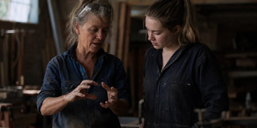 An experienced tradeswoman explaining her work to a younger apprentice at a workbench, illustrating how professional judgment passes from one generation to the next