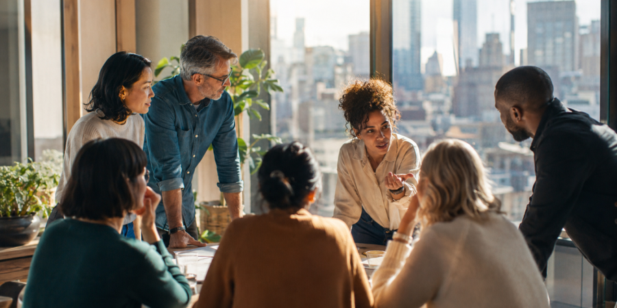 A bright, sunlit meeting room full of people in genuine conversation, illustrating shared decision-making under uncertainty, by Morris Misel